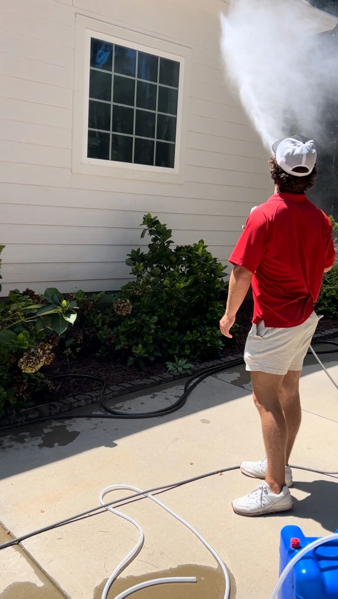 Person in red shirt pressure washing a house exterior with shrubs nearby.