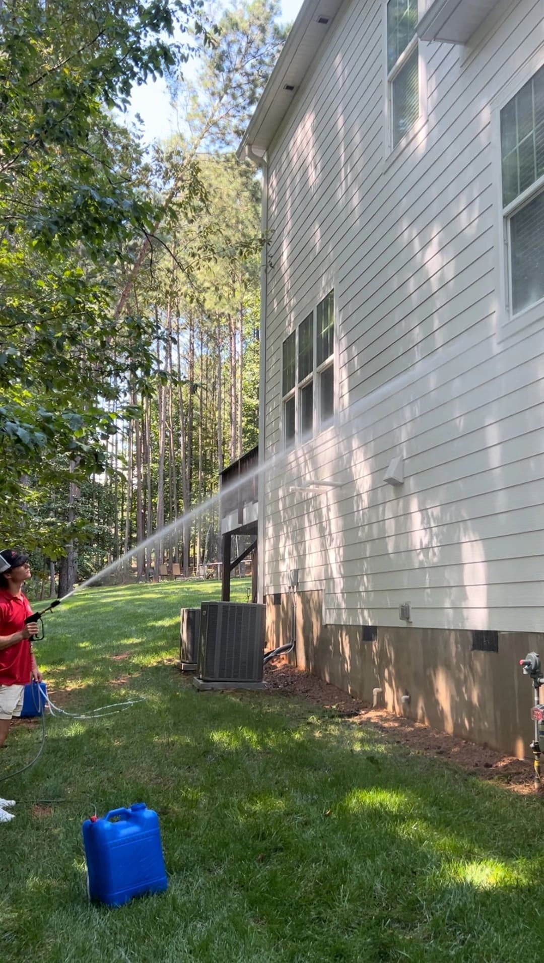 Man cleaning exterior of house with pressure washer near wooded area.