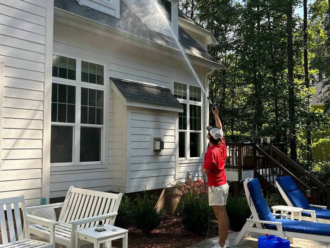 Woman power washing a house exterior with a spray nozzle, surrounded by patio furniture and greenery.