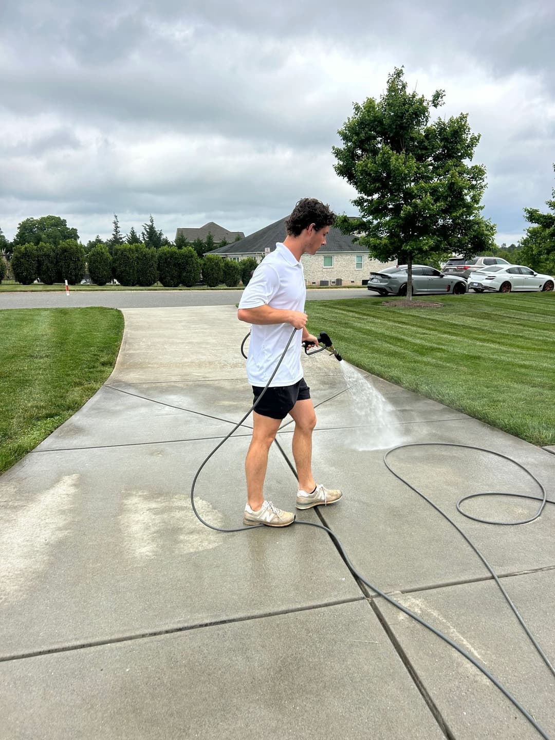 Person pressure washing a concrete driveway on a cloudy day, surrounded by neatly trimmed grass.