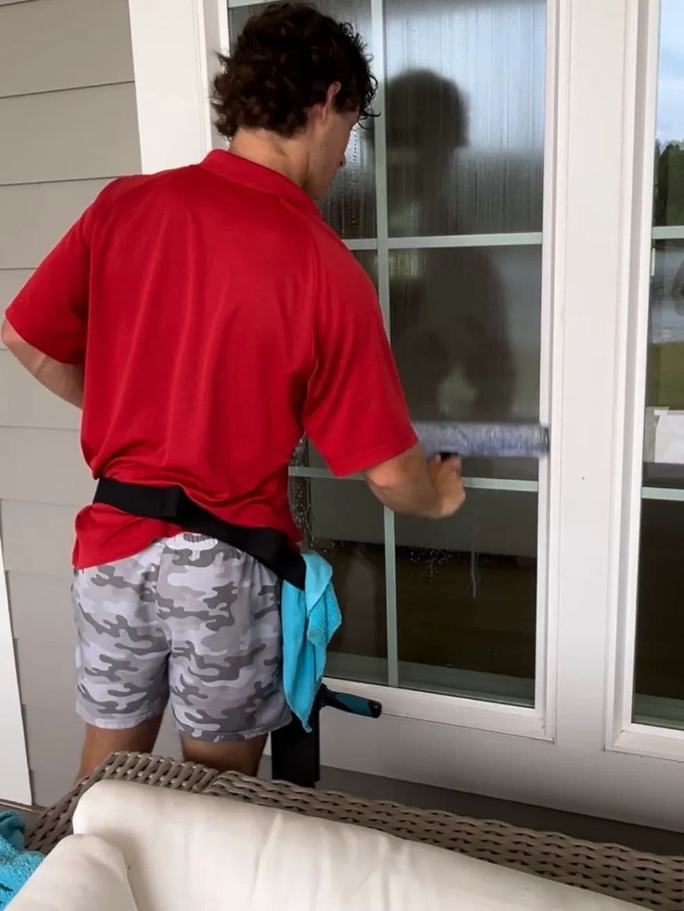 Man in red shirt and camo shorts cleaning glass door with a squeegee.