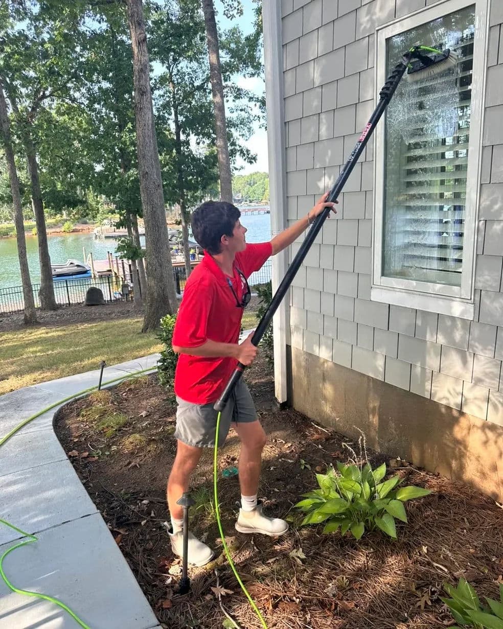 Person using a long pole to clean windows on a house near a lake surrounded by trees.
