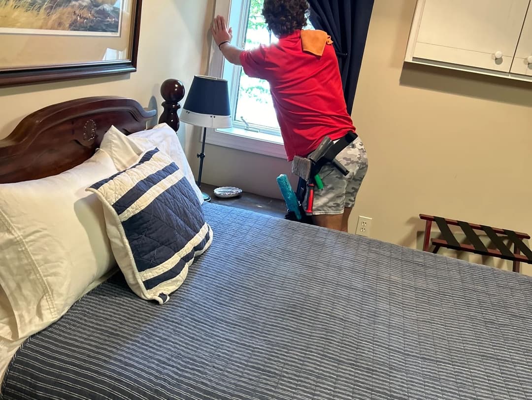 Person adjusting window blinds in a cozy bedroom with a neatly made bed and decor.