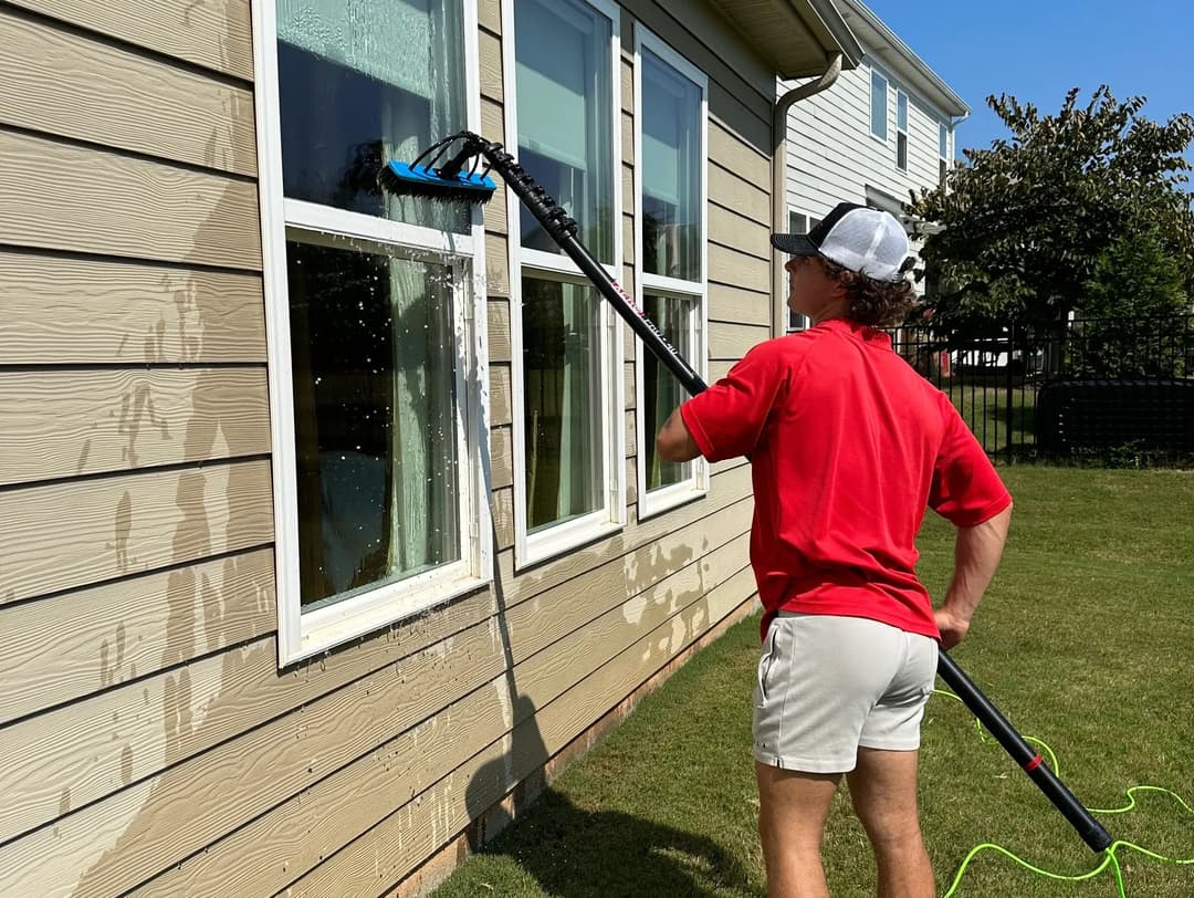 Person cleaning windows with a long squeegee tool on a sunny day outside a home.