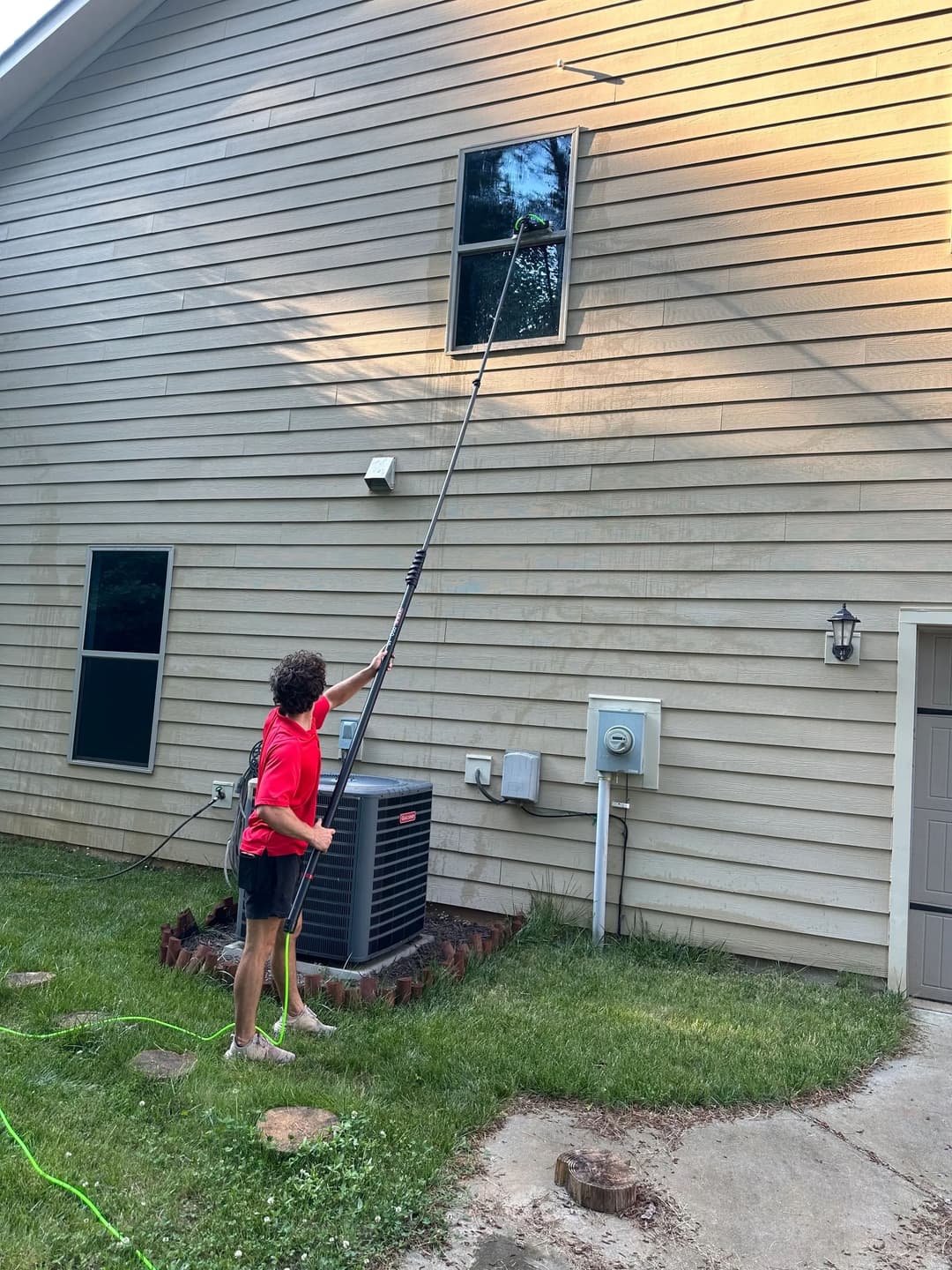 Person in red shirt cleaning a window on a two-story house using a long pole.
