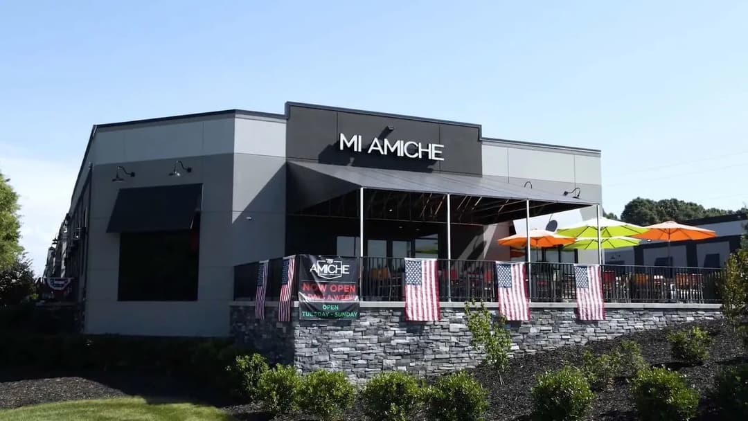 Exterior view of Mi Amiche restaurant with patio seating and American flags, now open.