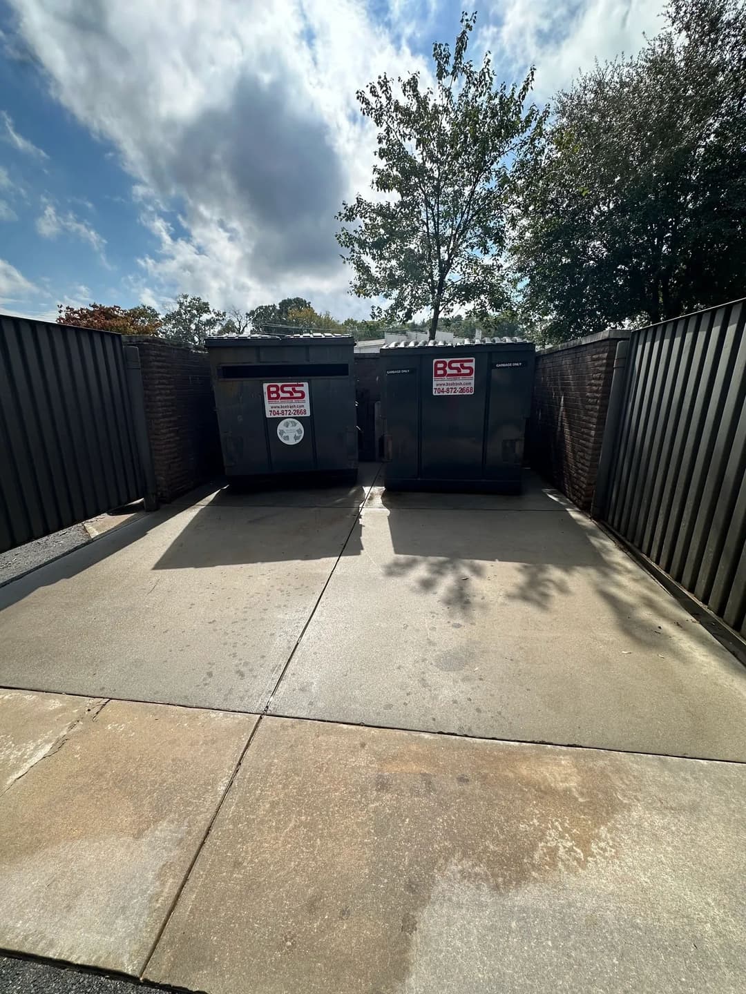 Two dumpsters in a fenced area, sunny day with clouds and trees in the background.
