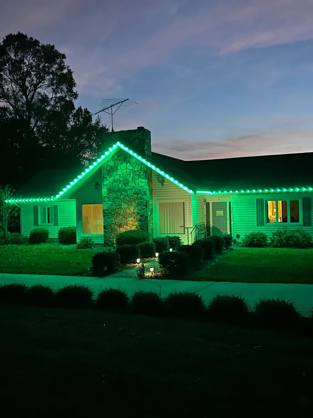 Green illuminated house at dusk with stone chimney and landscaped front yard.